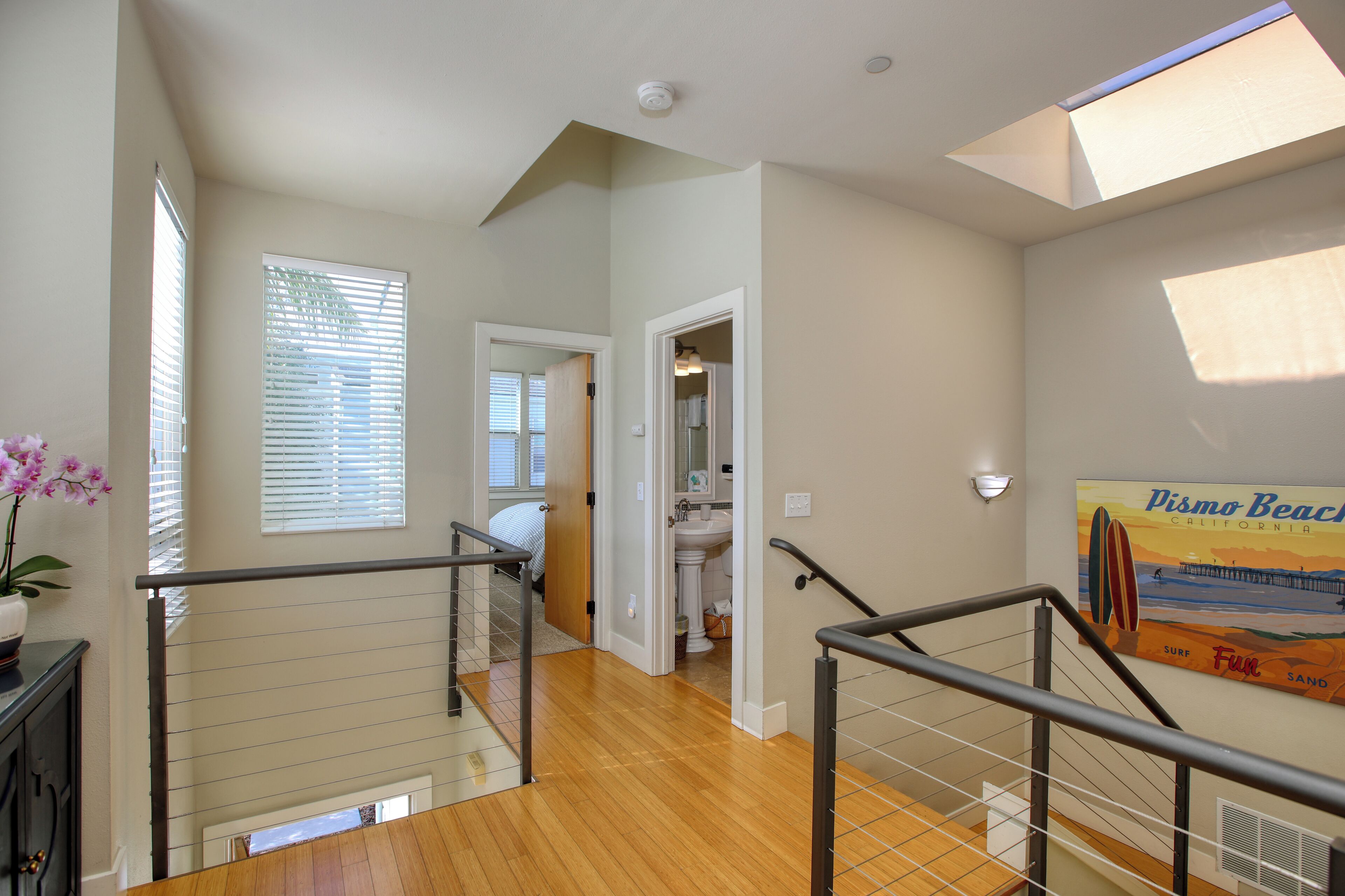 A spacious interior with a wooden staircase, a bathroom with a sink and mirror, and a poster of Pismo Beach.