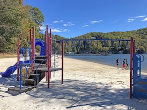 A playground with a slide and climbing structures, located on a sandy beach with a forested hill in the background.