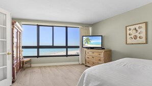 Habitación de hotel con un gran ventanal con vistas a una playa, una cama, un aparador de madera y un cuadro en la pared.