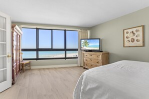 A hotel room with a large window overlooking a beach, a bed, a wooden dresser, and a framed picture on the wall.