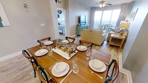Een eetgedeelte met een houten tafel voor vier personen, omringd door stoelen. Een televisie is aan de muur bevestigd, en op de achtergrond is een balkon met uitzicht te zien.