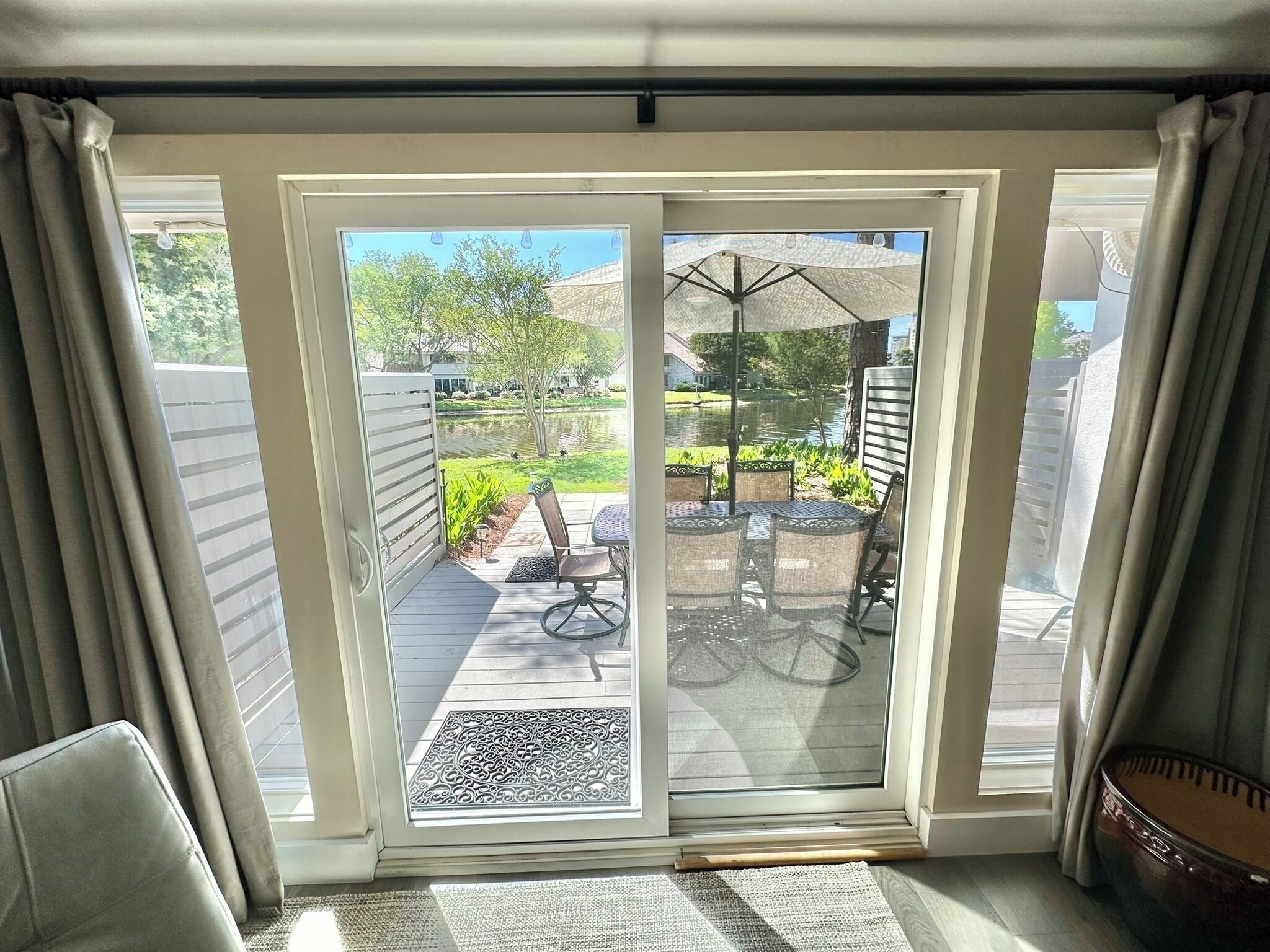 Une porte-fenêtre coulissante donne sur un patio extérieur aménagé avec une table et des chaises, un vélo, et offre une vue sur un étang et des arbres.