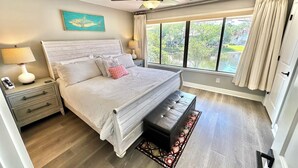 Un dormitorio con cama, mesitas de noche, ventilador de techo, ventana con cortinas y vistas al exterior con vegetación.
