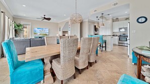 A modern dining area with a wooden table and woven chairs, a ceiling fan, and a kitchen in the background. - Lone Palm by GSVR | Charming Beachfront Retreat w/ Great Primary Bed/Bath + FREE Activities/Golf (Gulf Shores)