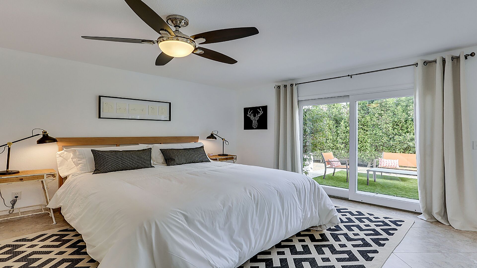 Een slaapkamer met een bed, een plafondventilator, een nachtkastje met een lamp, een ingelijst schilderij en uitzicht op een tuin door een schuifdeur.