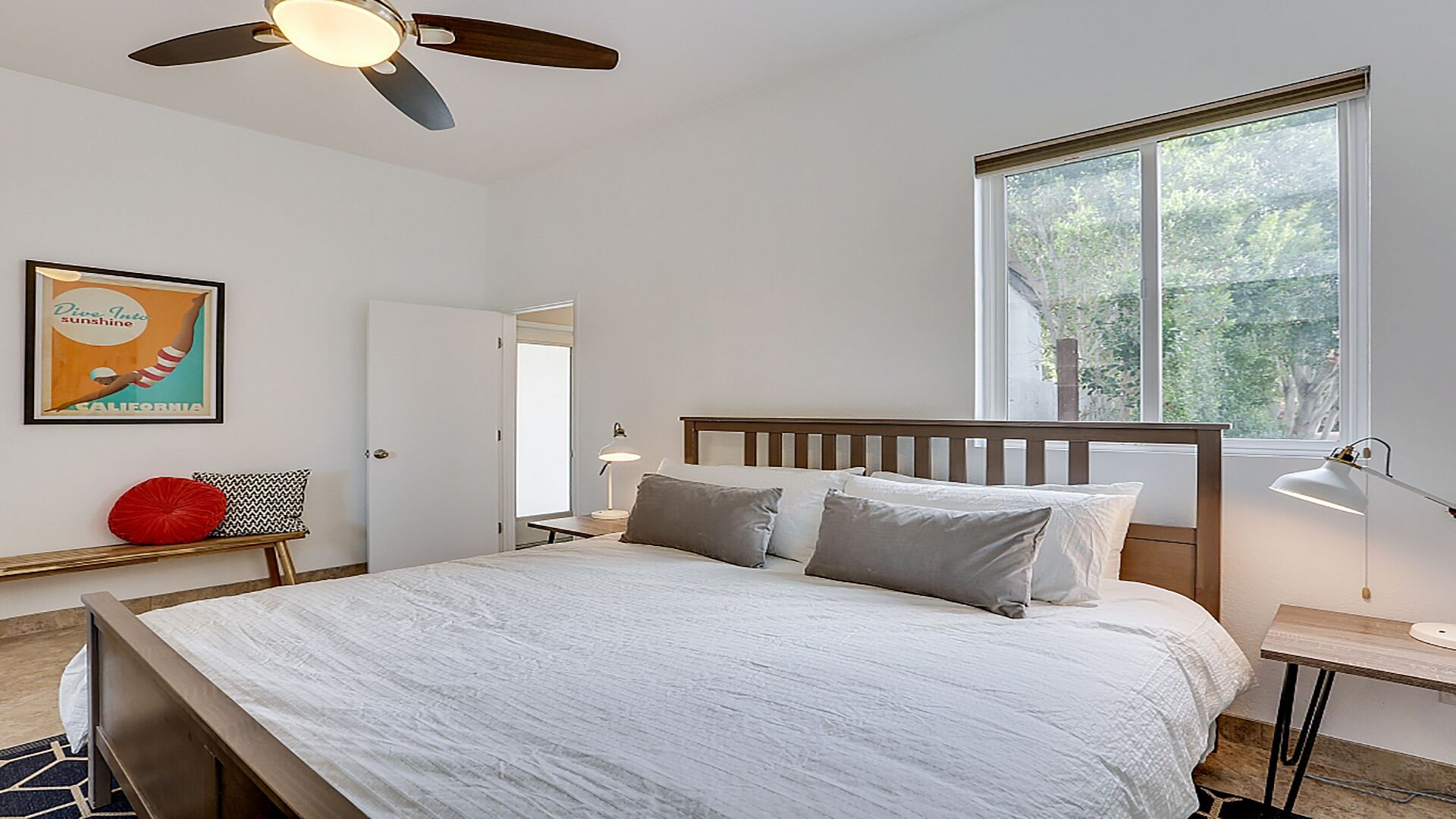 Ein ordentlich eingerichtetes Schlafzimmer mit Bett, Deckenventilator, Fenster mit Blick ins Grüne, Nachttisch mit Lampe und einem gerahmten Bild an der Wand.