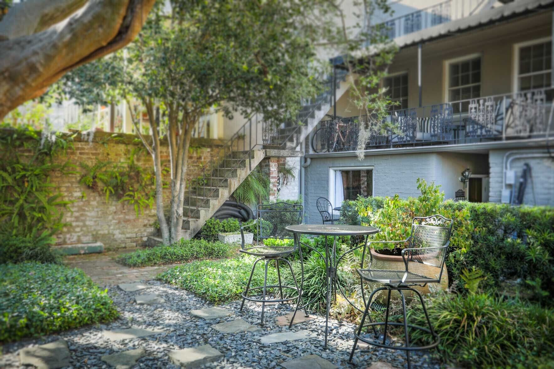 A courtyard with a stone pathway, metal chairs, and tables, surrounded by greenery and a building with a balcony.