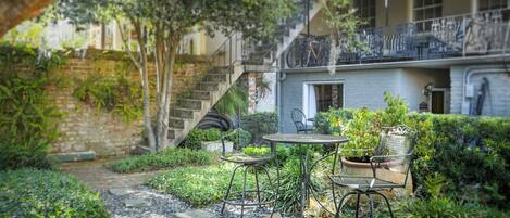 A courtyard with a stone pathway, metal chairs, and tables, surrounded by greenery and a building with a balcony.
