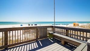 A wooden deck with a bench overlooking a beach with a small yellow building and beach chairs.