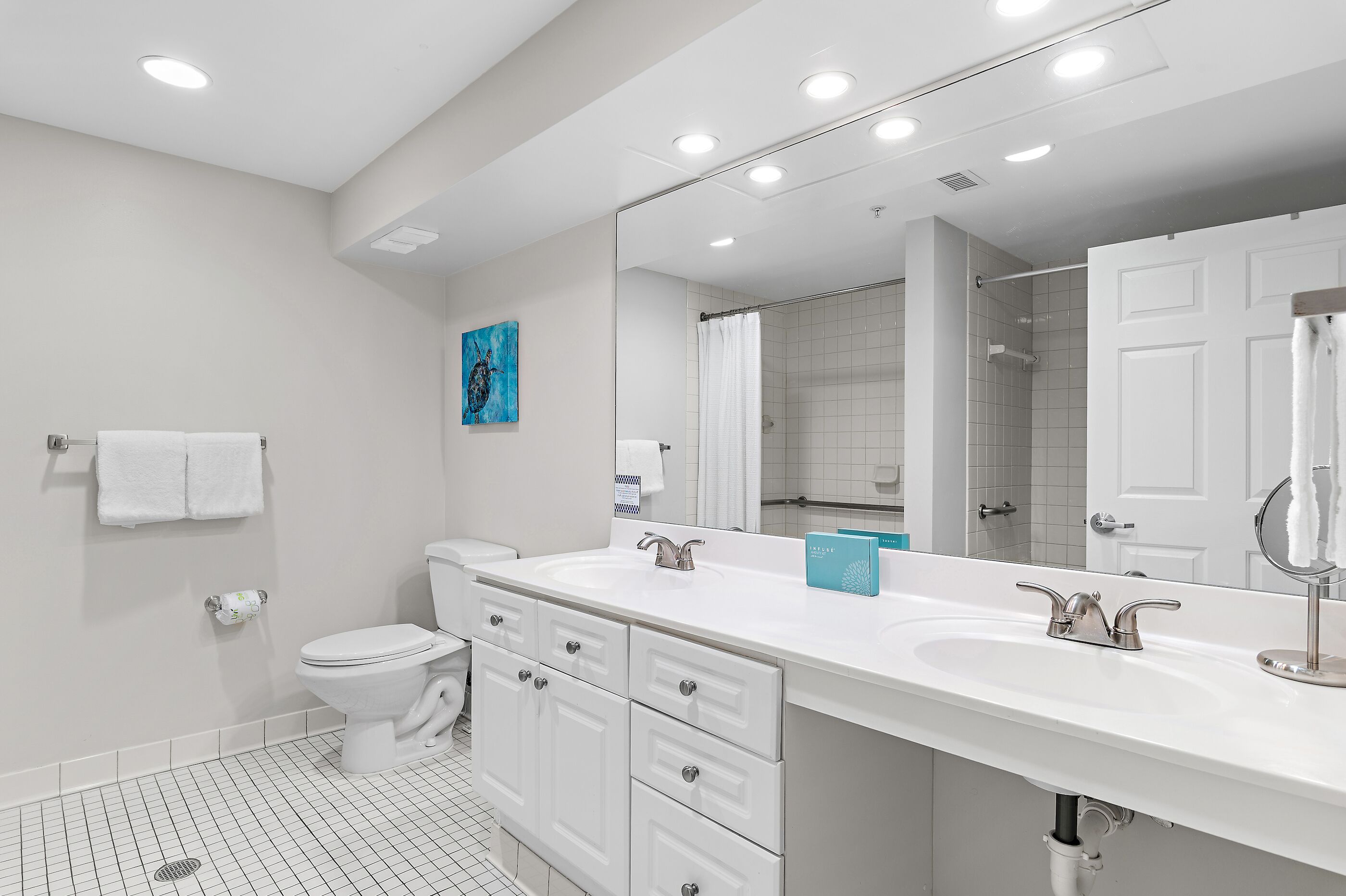Een moderne badkamer met een wit wastafelmeubel, een wastafel en een toilet. Boven de wastafel hangt een grote spiegel en er is een douchegedeelte zichtbaar op de achtergrond.