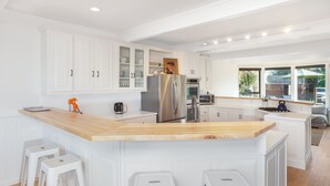 A modern kitchen with white cabinetry, a stainless steel refrigerator, a wooden island with bar stools, and a view of the outdoors through large windows. - Fresh and Lovely Inner Lagoon Home (Stinson Beach)
