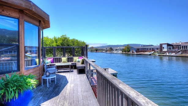 A wooden deck with outdoor seating, overlooking a calm river with boats and buildings in the background. - Fresh and Lovely Inner Lagoon Home (Stinson Beach)