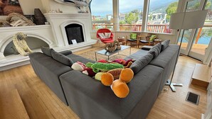 A modern living room with a large gray sectional sofa, a fireplace, and a view of the ocean through large windows. - Fresh and Lovely Inner Lagoon Home (Stinson Beach)