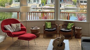 A spacious living room with large windows, a red chair, a wooden coffee table, and a view of a lake and mountains. - Fresh and Lovely Inner Lagoon Home (Stinson Beach)