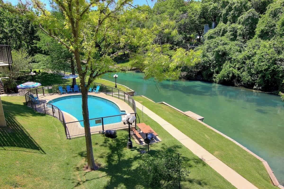 Una piscina con muelle de madera, rodeada de césped y árboles, con vista a un río.