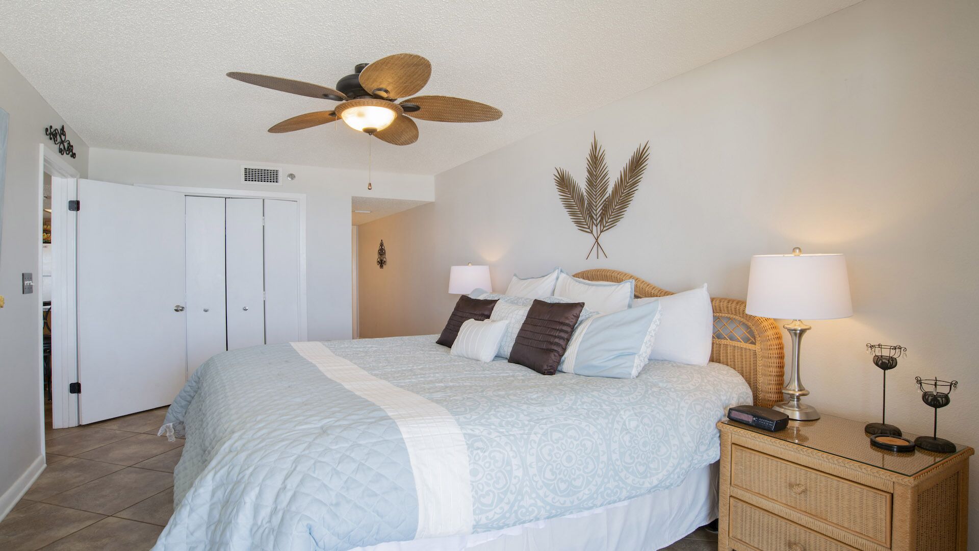 A bedroom with a bed, a ceiling fan, two bedside lamps, a decorative wall piece, and a wicker nightstand.