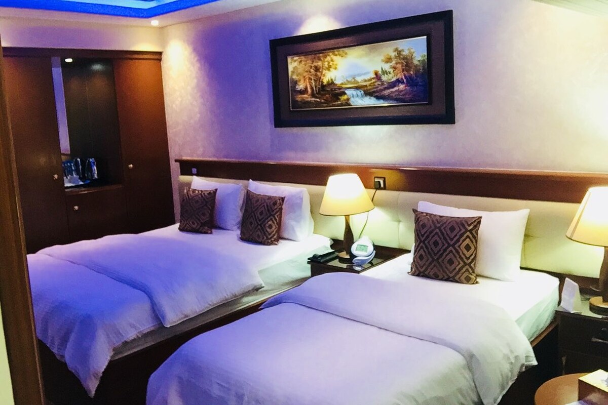 A hotel room with two beds, a painting on the wall, and a ceiling with blue lighting.