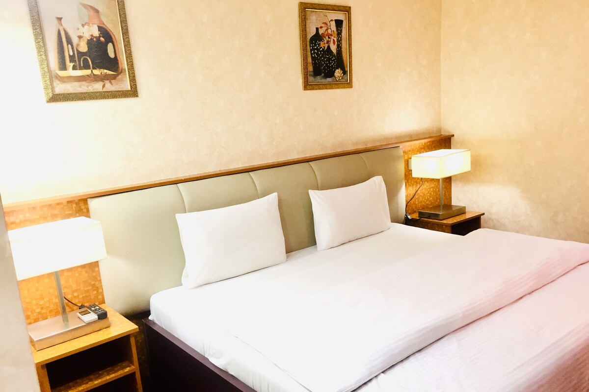 A hotel room with a large bed, two bedside tables, a nightstand, a lamp, and two framed pictures on the wall.