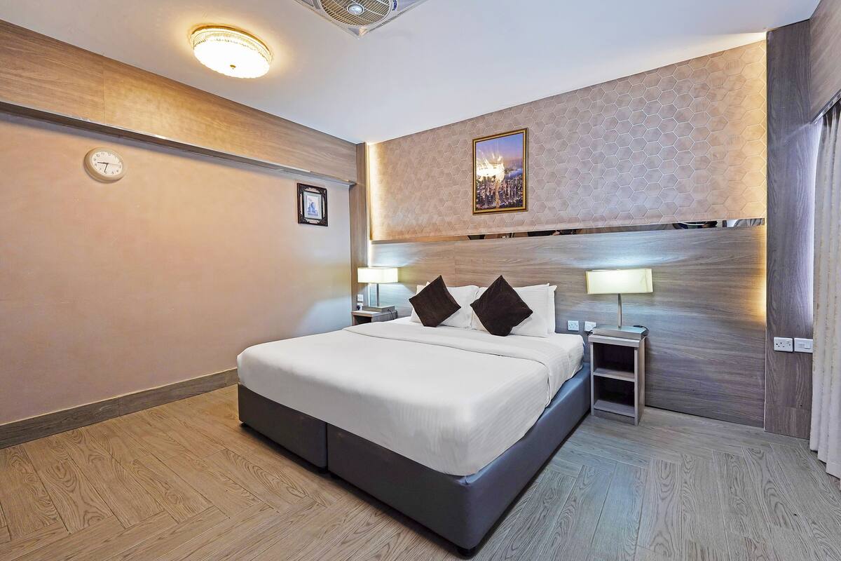 A modern hotel room with a large bed, bedside tables with lamps, a clock on the wall, and a cityscape artwork above the bed.