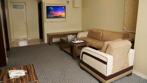 Een hotelkamer met een beige bank, een houten bijzettafeltje, een flatscreen televisie en een telefoon op een kamerbreed tapijt met dessin.