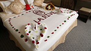 Een netjes opgemaakt bed met een hartvormige bloembladdecoratie van rozen, een handdoek, een teddybeer en een bloemenboeket.