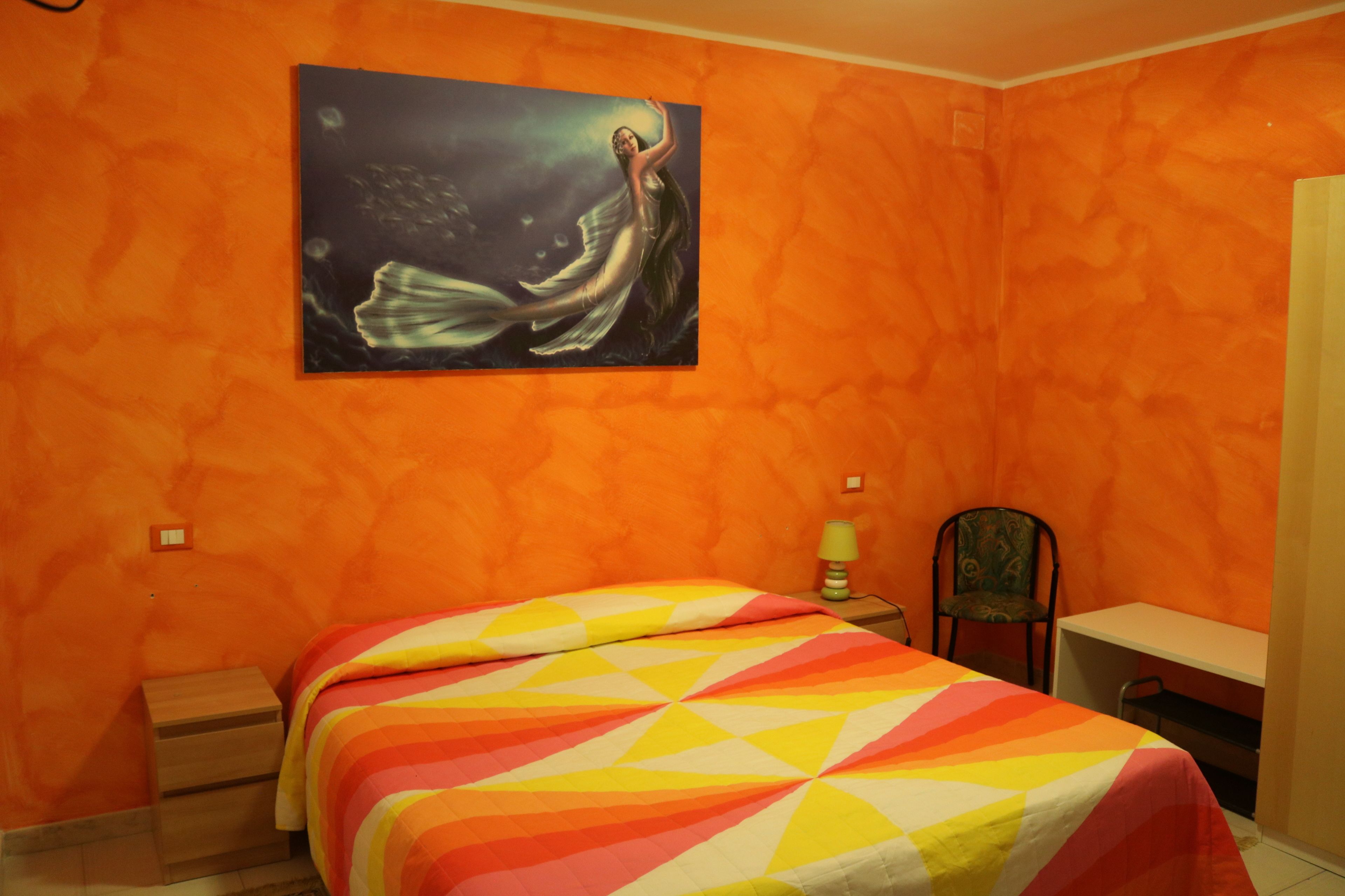 A room with orange walls, a bed with a colorful striped bedspread, a desk, a chair, and a painting of a woman on the wall.