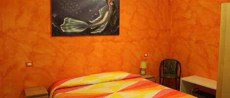 A room with orange walls, a bed with a colorful striped bedspread, a desk, a chair, and a painting of a woman on the wall.