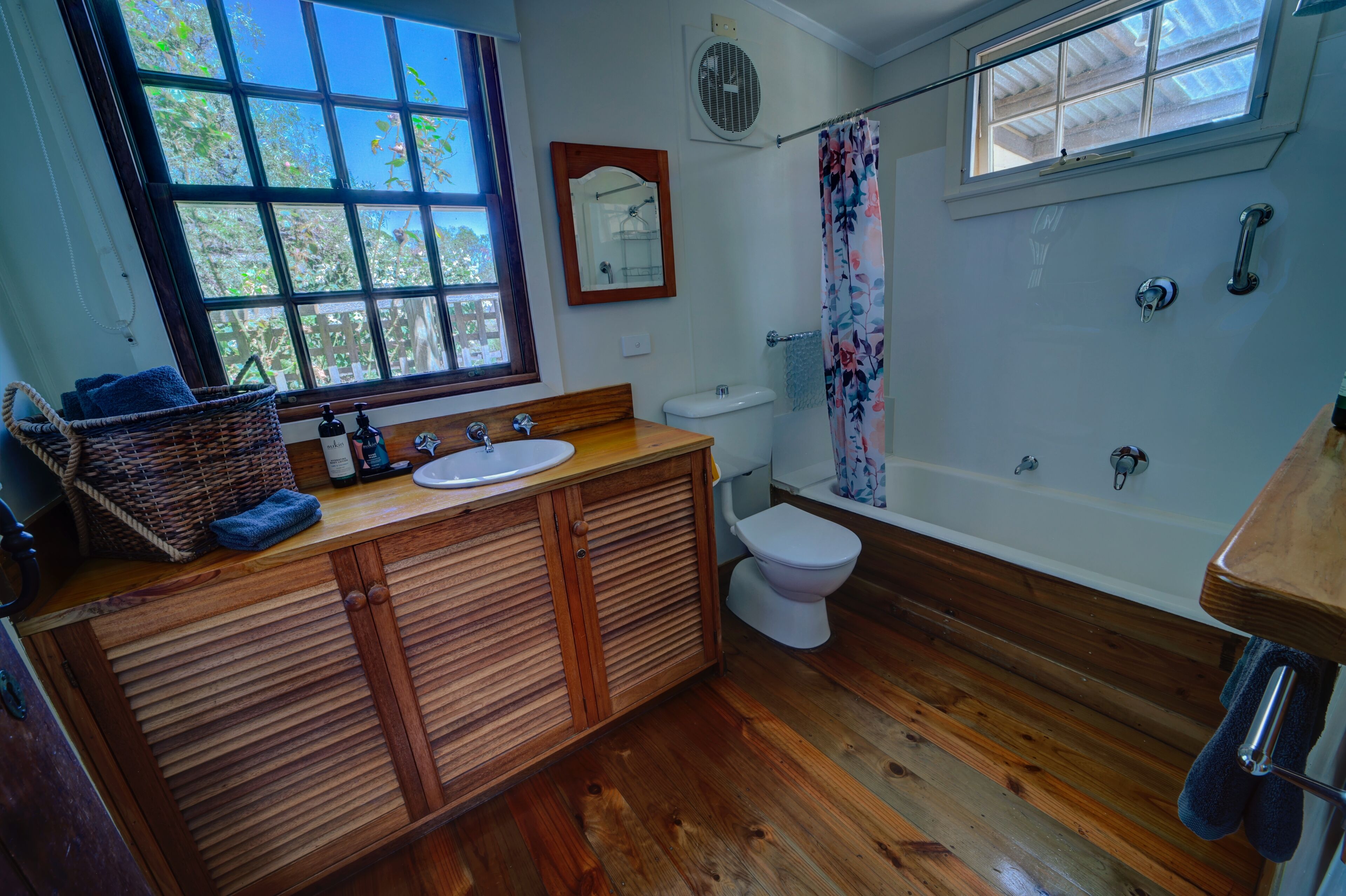 Een badkamer met houten vloeren, een houten wastafelmeubel met een wastafel, een toilet, een douche met een bloemen gordijn en een raam met uitzicht op bomen.
