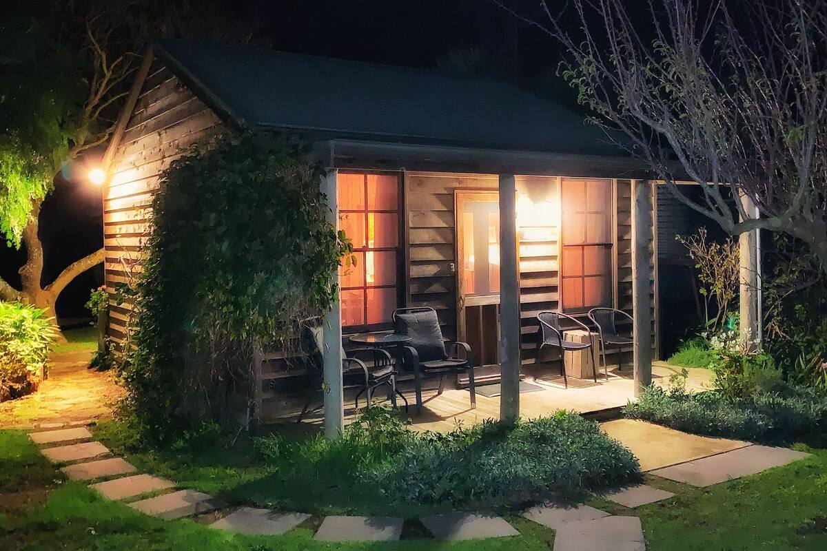 A cozy outdoor seating area with a wooden table and chairs, surrounded by greenery and a stone pathway leading to a wooden building with a lit interior.
