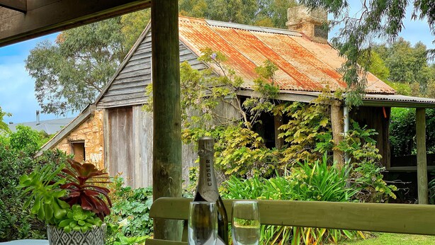 A bottle of CROSER wine and a glass on a table with a patterned tablecloth, set outdoors with a view of a rustic building and lush greenery. - Cherry Plum Cottages (Port Fairy)
