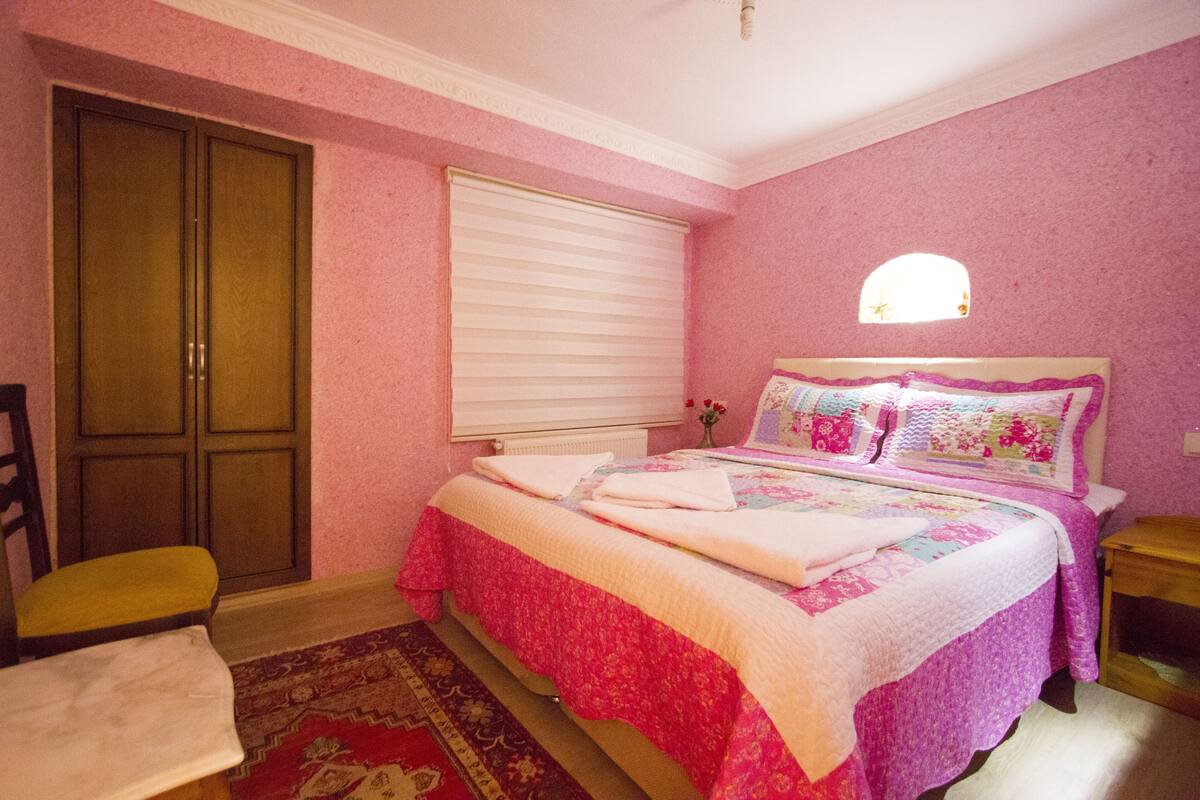 Ein Schlafzimmer mit rosa Wänden, einem Bett mit floralem Bettbezug, einem Holzschrank, einem Stuhl und einem kleinen Tisch.