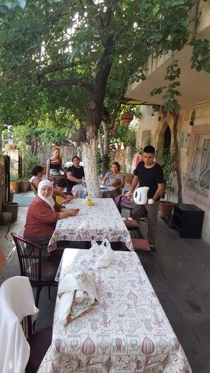 A group of people seated at a table with a patterned tablecloth, under a tree, in an outdoor setting with a building in the background. - Nil Story House (Nevsehir)