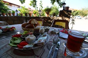Une table dressée à l’extérieur, avec une variété d’aliments, notamment du pain, des légumes et un verre de thé, des plantes en pot et un mur de pierre en arrière-plan.