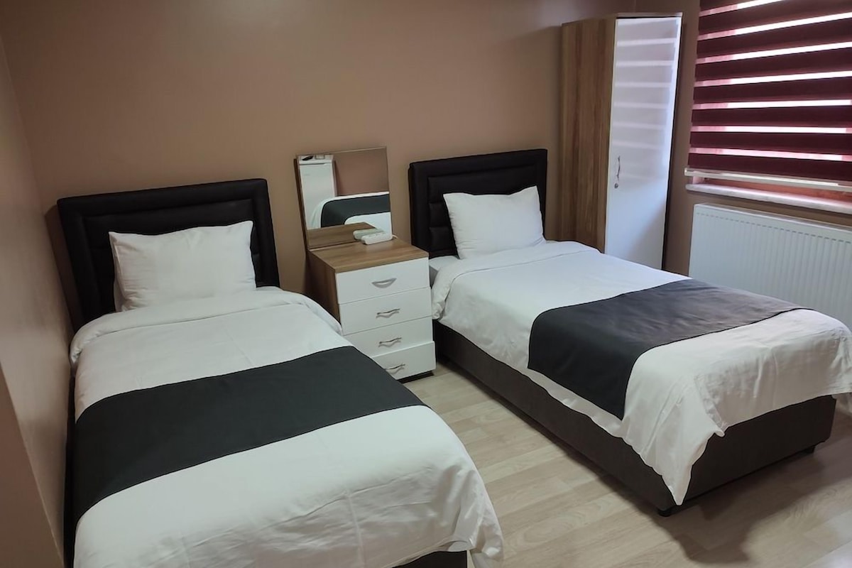 A hotel room with two beds, a bedside table, a mirror, and a window with blinds.