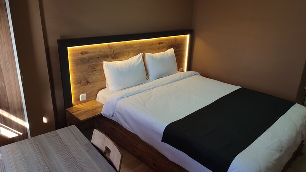 Un letto rifatto con cura, con lenzuola bianche e una coperta nera, una testiera in legno e un comodino con una lampada.