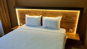 Un letto rifatto con lenzuola e cuscini bianchi, una testiera in legno con illuminazione integrata e un piccolo comodino con presa di corrente.