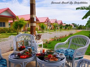 A dining area with wicker chairs and a table set with breakfast items, overlooking a well-maintained garden and a building with red roofs. - Sermsuk Resort (Pranburi)