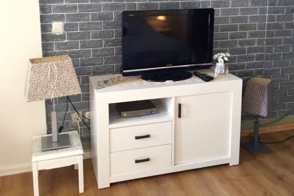 A room with a brick wall, a white entertainment unit with a flat-screen TV, a small table with a lamp, and a flower vase.