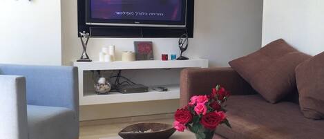 A living room with a flat-screen TV mounted on the wall, a blue armchair, a brown sofa, a coffee table with a vase of flowers and magazines, and a white shelf with decorative items.