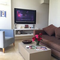 Un salón con televisión de pantalla plana montada en la pared, un sillón azul, un sofá marrón, una mesa de centro con un jarrón de flores y revistas, y un estante blanco con adornos.