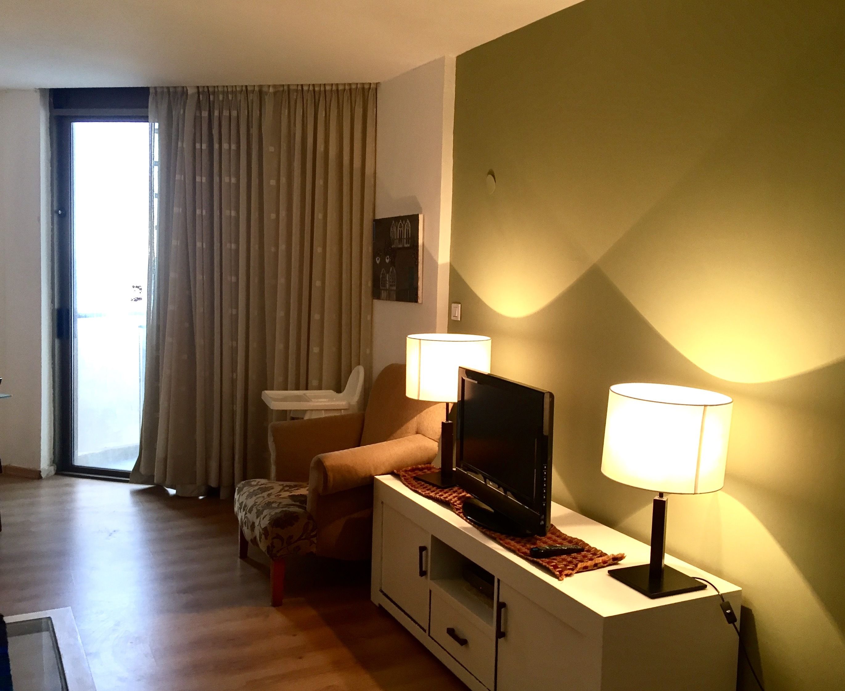 Uma sala de estar com um sofá, uma televisão sobre um móvel, duas luminárias, uma janela com cortinas e uma pequena mesa com um controle remoto.