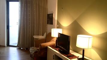 Uma sala de estar com um sofá, uma televisão sobre um móvel, duas luminárias, uma janela com cortinas e uma pequena mesa com um controle remoto.