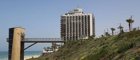 Un paseo marítimo con un edificio moderno al fondo, un puente y una playa con palmeras.