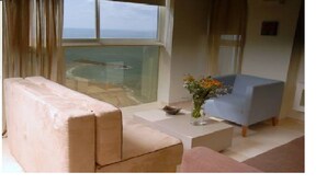 A living room with a beige sofa, a blue armchair, and a small table with a vase of flowers. A large window offers a view of the ocean. - Raphael Apartments (Herzliya)