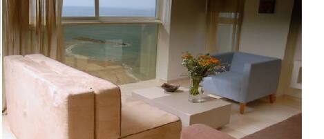 A living room with a beige sofa, a blue armchair, and a small table with a vase of flowers. A large window offers a view of the ocean.