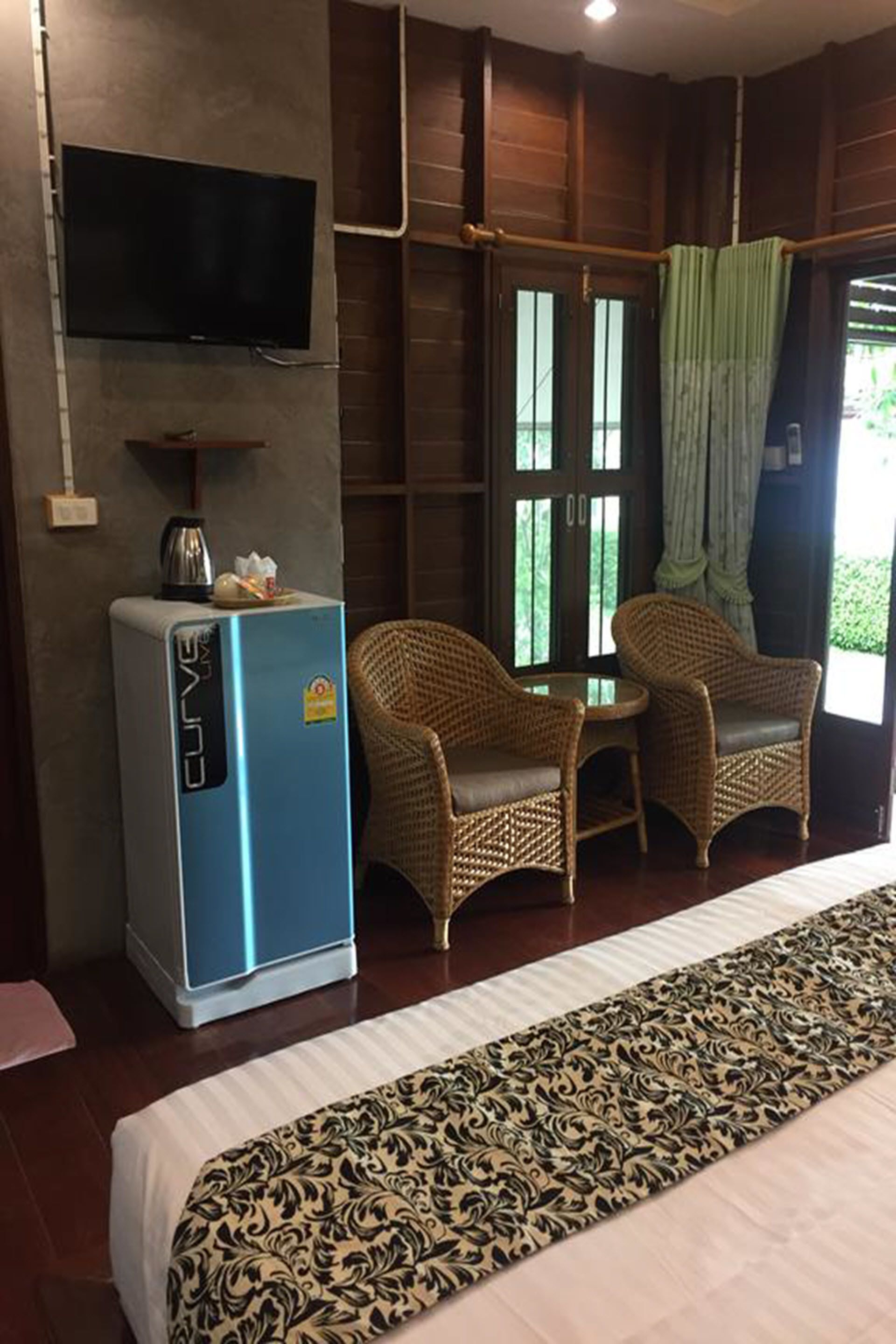 A hotel room with a bed, a television mounted on the wall, a refrigerator, two wicker chairs, and a window with curtains.