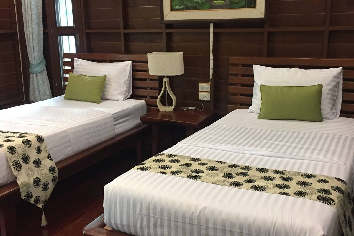 A hotel room with two beds, wooden headboards, a painting of a garden, a bedside table with a lamp, and a window with curtains.