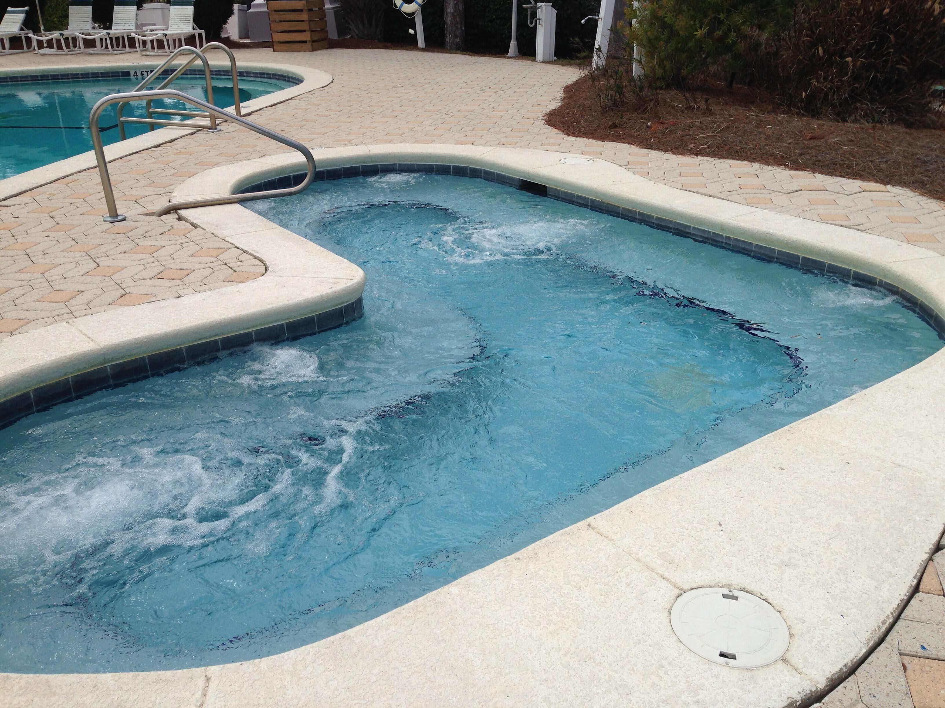 Une piscine avec un petit espace jacuzzi, entourée d’une terrasse dallée et de chaises longues.