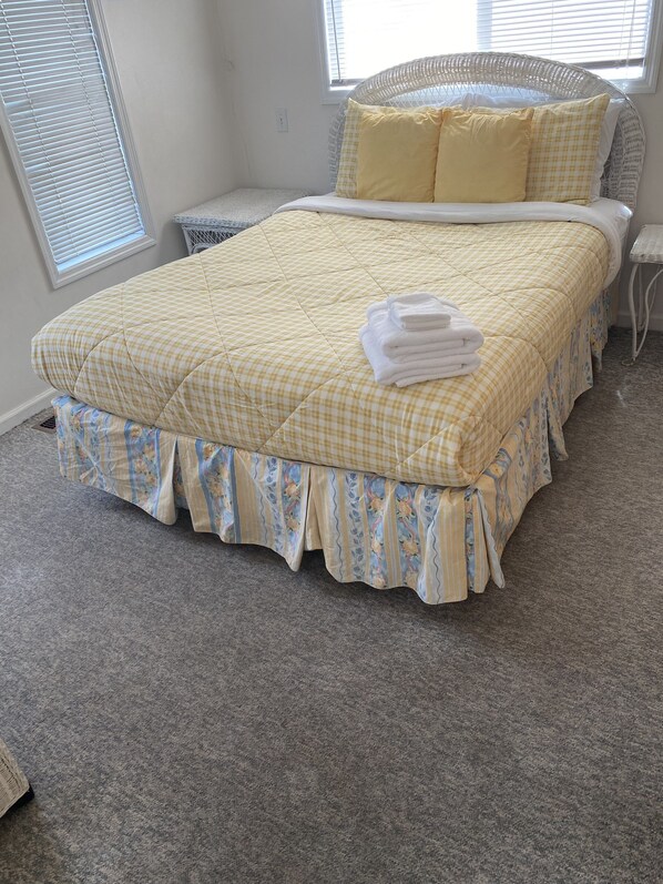 A neatly made bed with a yellow checkered bedspread and matching pillowcases, a white wicker headboard, and a small bedside table with a lamp.