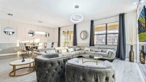 Ein modernes Wohnzimmer mit Ecksofa, runden Hocker, einer Essecke und großen Fenstern.
